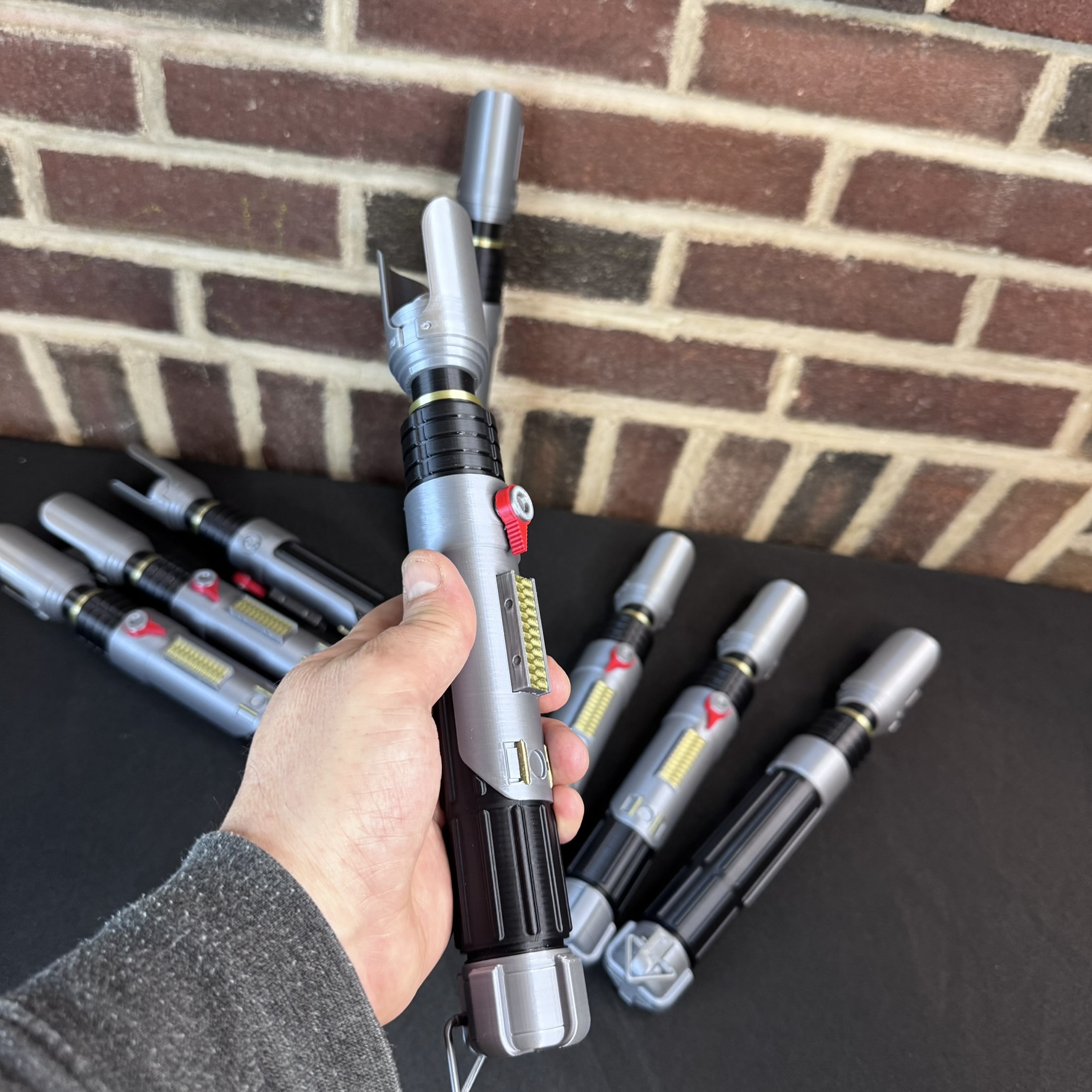 Hand holding a Sabine Wren lightsaber prop hilt in front of several additional hilts displayed on a table.