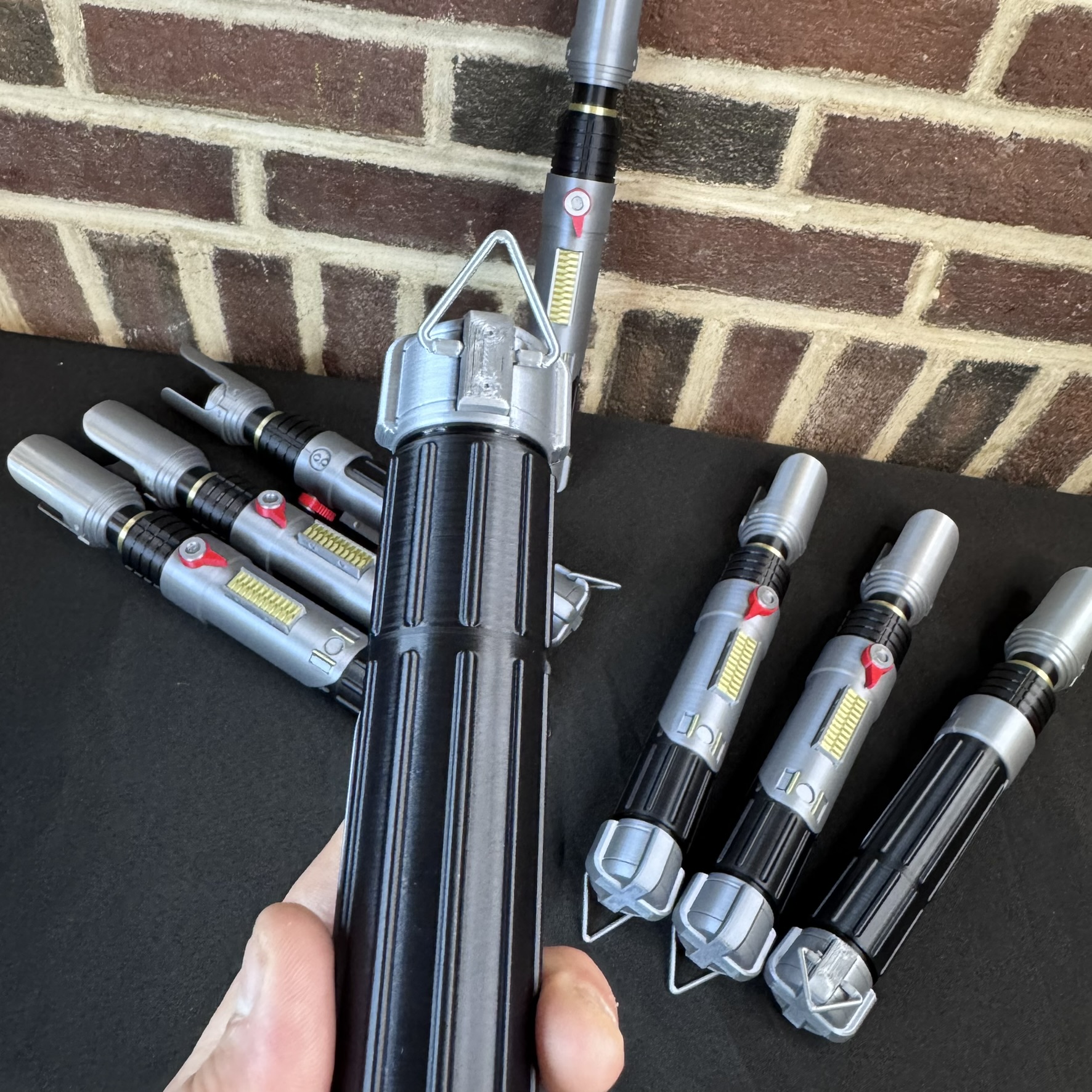 Hand holding a Sabine Wren lightsaber prop hilt in front of several additional hilts displayed on a table.